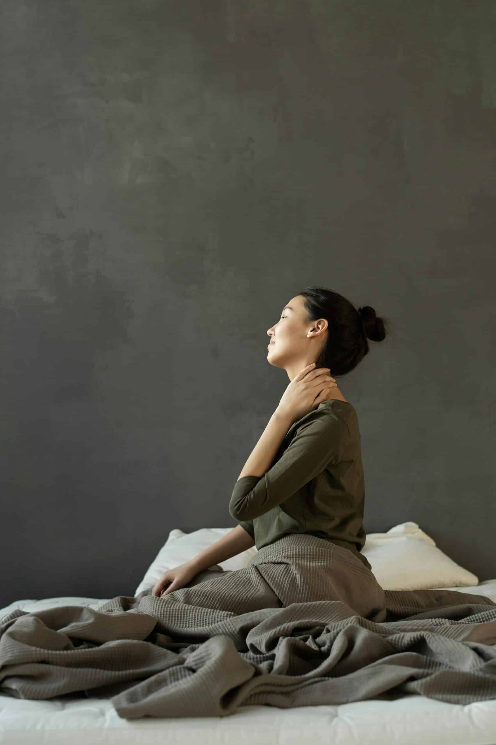 Vrouw zit rechtop in bed met hand op haar nek, mogelijk bij pijn, tijdens een moment van bewustwording of oefening uit Mindfulness-Based Pain Management.